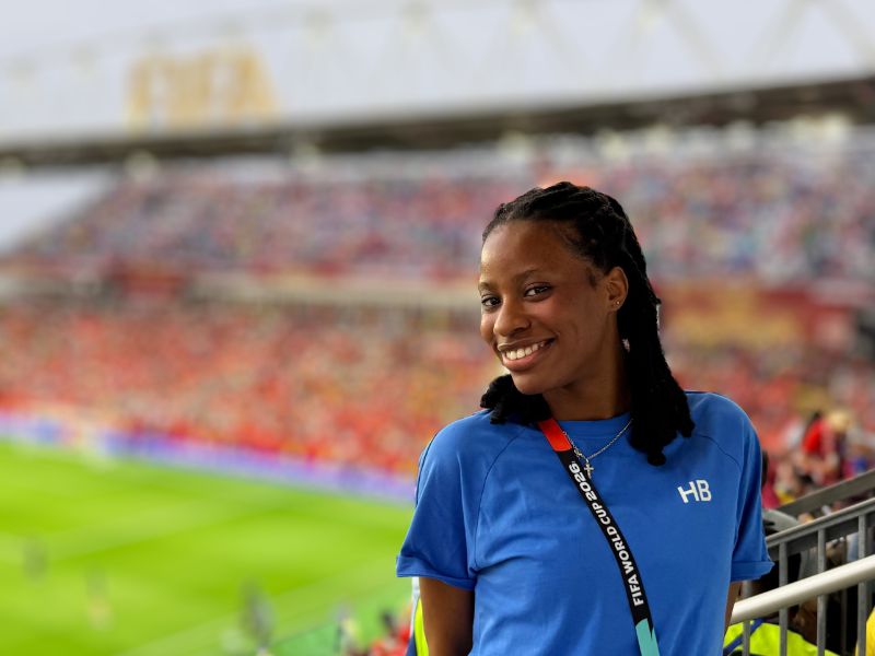 Jessica Edet-Nankere smiles from a seating area overlooking the pitch at Toronto Stadium during a FIFA World Cup 2026 match. Wearing a blue FIFA Host Broadcast (HB) Intern Programme shirt and accreditation, she stands in front of a packed crowd with the field and FIFA World Cup branding visible in the background.