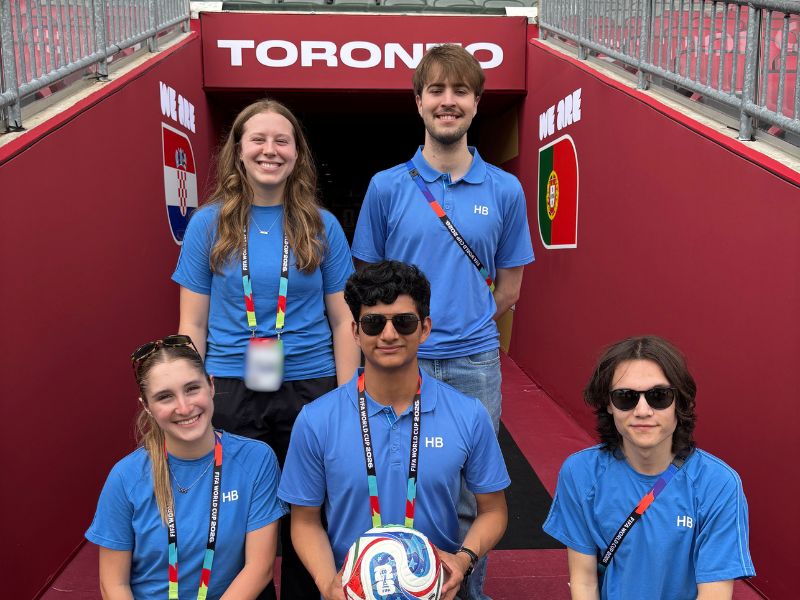 Five Toronto Metropolitan University Media Production students wearing blue FIFA Host Broadcast (HB) Intern Programme shirts and accreditation pose in the player tunnel at Toronto Stadium during the FIFA World Cup 2026. Marvin Aguilar-Martinez holds a FIFA match ball while the group smiles in front of the tunnel entrance, with the words "TORONTO" displayed above them.
