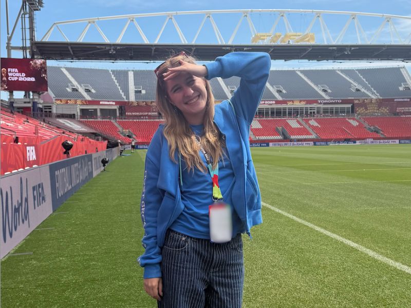  Keeley Pepper smiles while standing on the field at Toronto Stadium during the FIFA World Cup 2026. Wearing a blue jacket and FIFA accreditation, she poses near the touchline with the stadium seating, tournament branding and soccer pitch visible in the background.