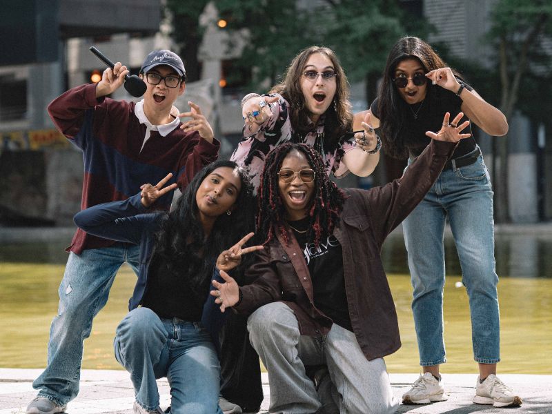 Five Student Communications content creators pose together outdoors on the TMU campus. Ria Sidhu stands at the back right, wearing sunglasses while smiling with her teammates, who are making playful poses for the camera in front of Kerr Hall and campus greenery.
