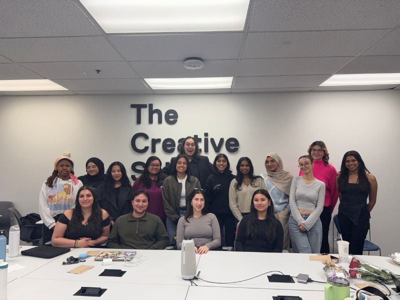 Members of The Society of The Creative School pose together in a meeting room beneath The Creative School wall sign. Ria Sidhu stands in the centre alongside fellow student leaders, while other members are seated around a conference table following a society meeting.