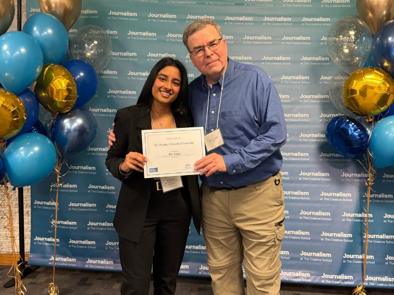 Ria Sidhu holds her Weather Network Scholarship certificate while posing with Journalism professor Gary Gould in front of a School of Journalism backdrop decorated with blue and gold balloons.