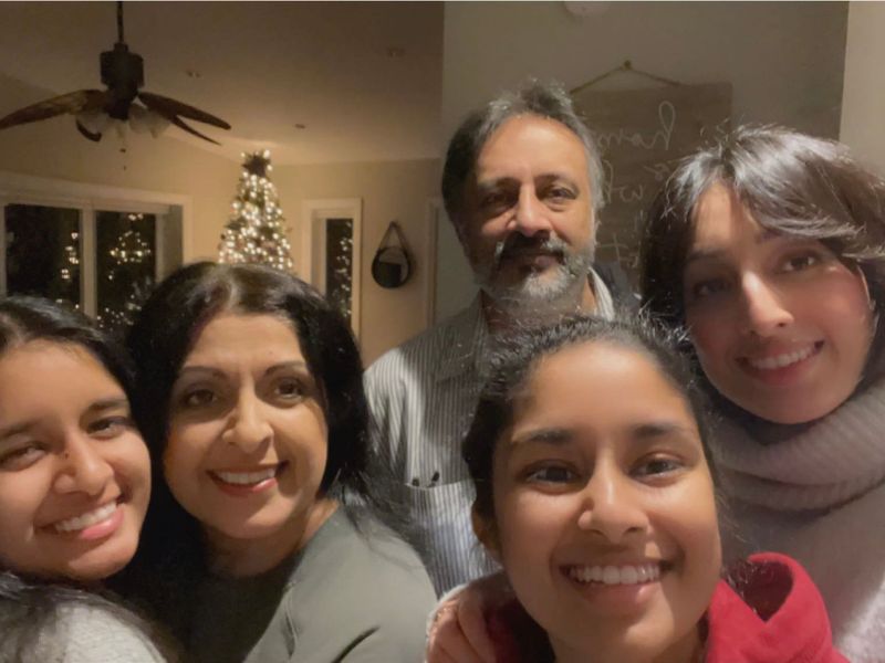 Ria Sidhu smiles alongside four family members in a selfie taken inside their home. A decorated Christmas tree is visible in the background as the family poses closely together for the photo.