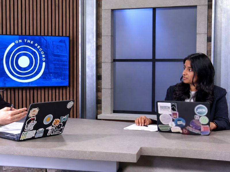 Ria Sidhu sits at a news desk in Studio D at the Rogers Communications Centre during a Journalism class, anchoring an episode of On the Record, TMU's student-produced news program. Another student sits across from her with laptops open in the broadcast studio.