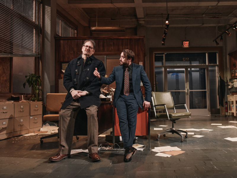 Two men perform on a messy office-themed stage; one sits on a desk while the other gestures mid-conversation.