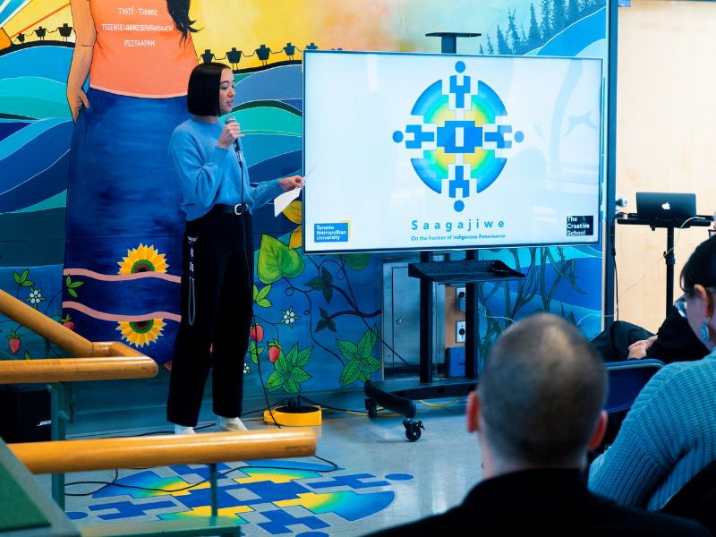  A woman stands beside a television screen speaking to a group of people sitting down.