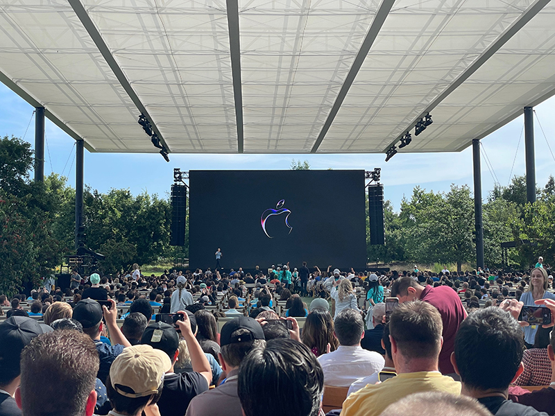 A crowd is gathered on seats in front of an outdoor stage. A man is on the stage with a microphone in hand. The apple logo is displayed on a screen behind him.
