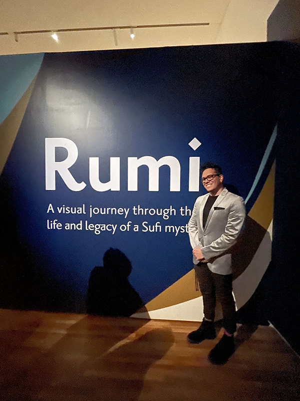 Carter-Arlt is standing in front of a wall with “Rumi A visual journey through the life and Legacy of a Sufi mystic” written on it 