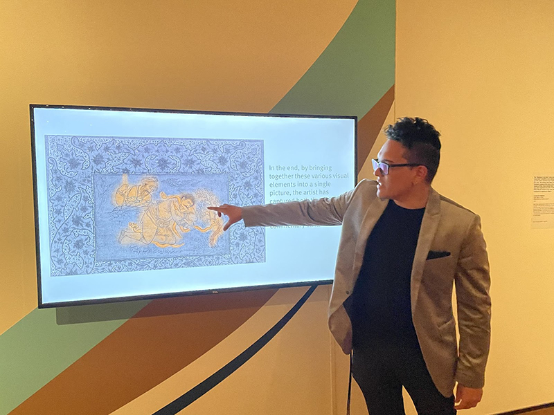 Carter-Arlt points to a highlighted section of a folio from an ancient manuscript that is displayed on a large TV monitor on a wall