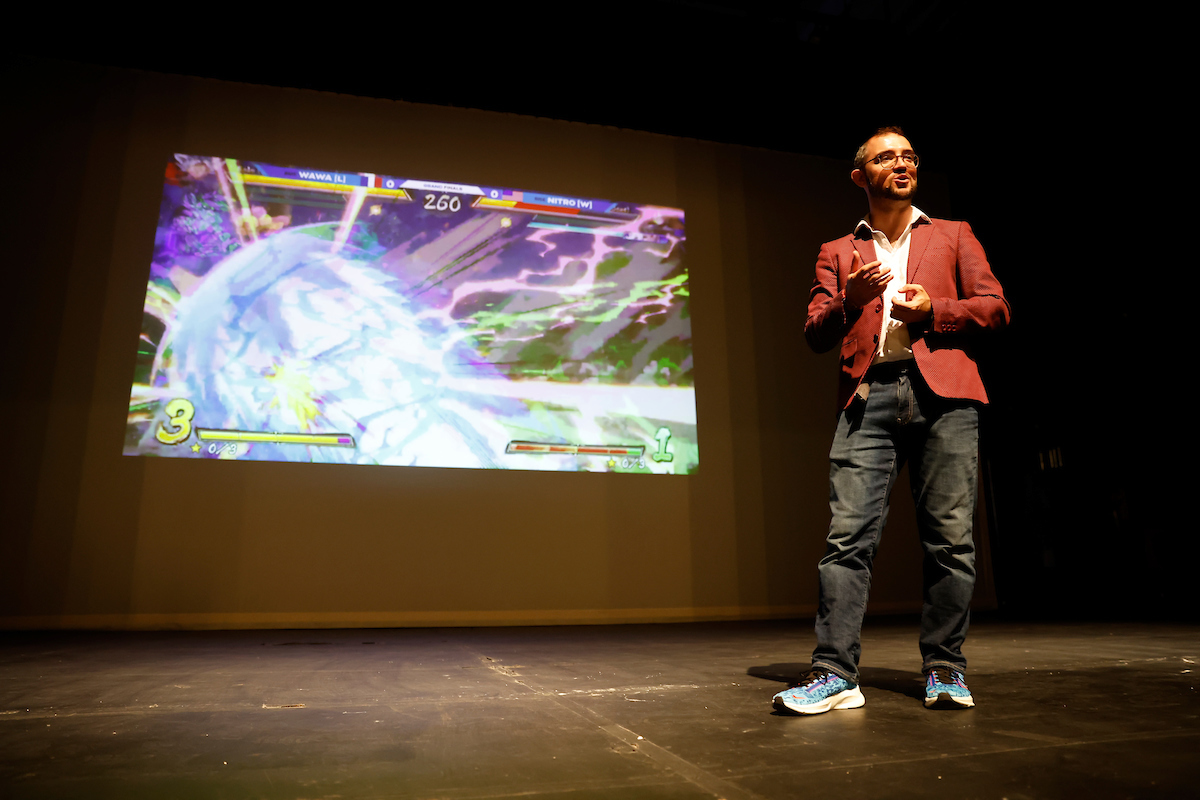 Geoffrey Lachapelle is speaking on stage of TMU’s Theatre in front of a backdrop showing a video game scene.