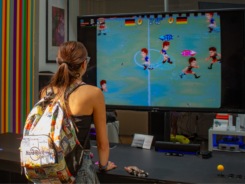 Two students stand in front of an arcade table, directing their attention to a digital video screen.