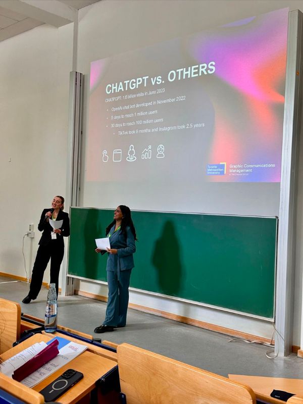 Two people on stage talking with a projecter screen in the background presenting the title ChatGPT vs. Others