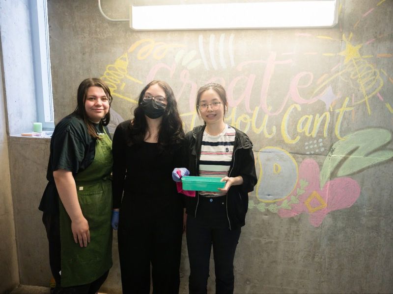 Three students facing and smiling to the camera in front of a concrete wall with colorful chalk drawings