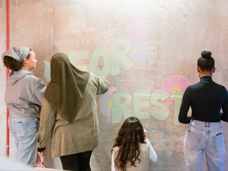 Four students using colorful chalk to draw an inspirational quote and cute flower drawings on a concrete wall.