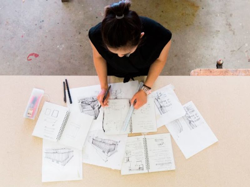 An overhead shot of a women working at a table with multiple sheets of paper and pencils on it