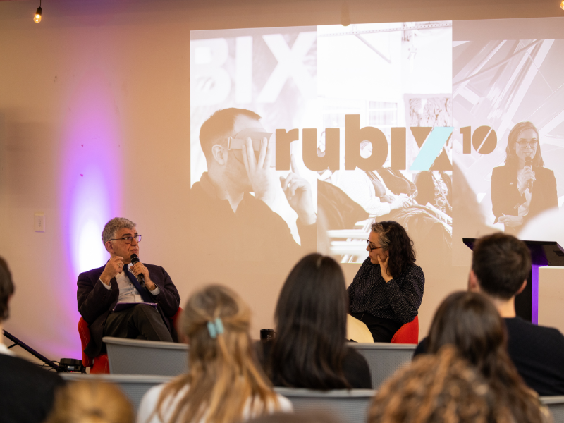 A man and a woman sitting down with mic in their hands talking, and audience is in front of them 