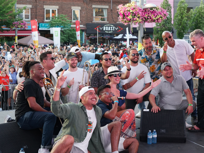 Medium-long shot of 13 people on an outdoor stage posing for a photo with a crowd of people in the audience behind them.