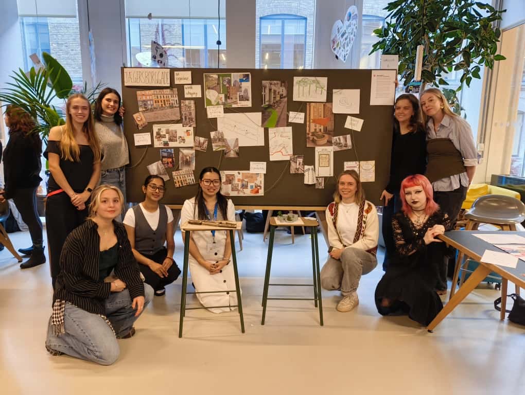 A group of students posing beside a presentation board filled with sketches, diagrams, and design ideas during the International Design Workshop at EK – Business Academy Copenhagen.