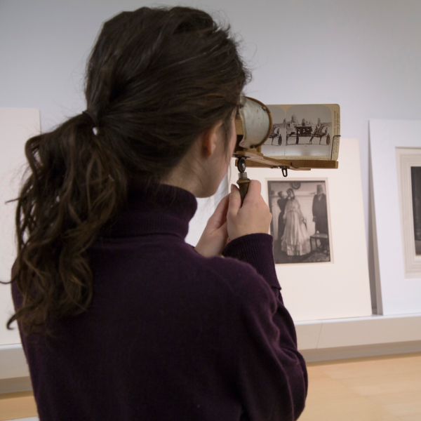 Medium close-up shot of the back of a woman's head as she holds a maginifying glass and looks at a wall of artifacts.