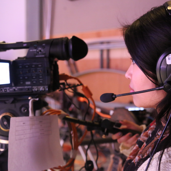 Close-up side shot of a woman wearing a mic headset and standing behind a recording camera.