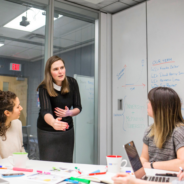 Three people are talking abd gathered around a table with writing utensils. One is standing and explaining something with a small futuristic white robot next to her. There is a whiteboard wall in the back with writing on it. 