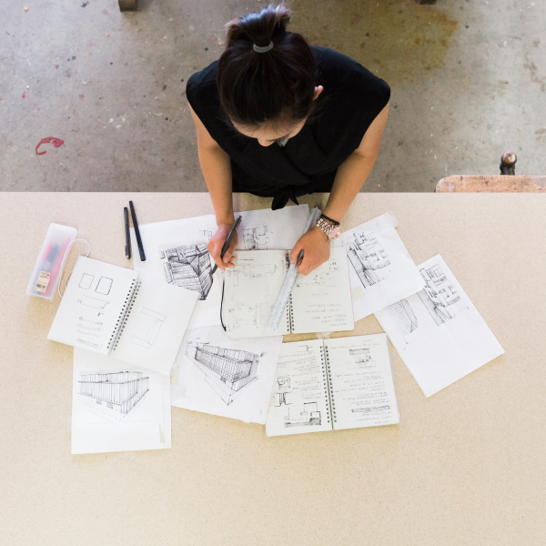 Aerial wide shot of a woman looking down at a table with several pieces of paper with sketches, floor plans, and other work.