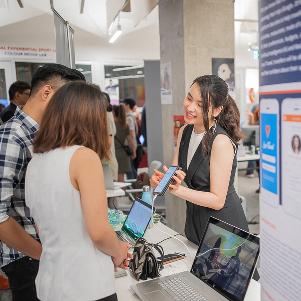 Medium-long shot of a woman showing something on her phone to two people. There is at table between them with two laptops in it. She is presenting something and is at an event with attendees in the background.