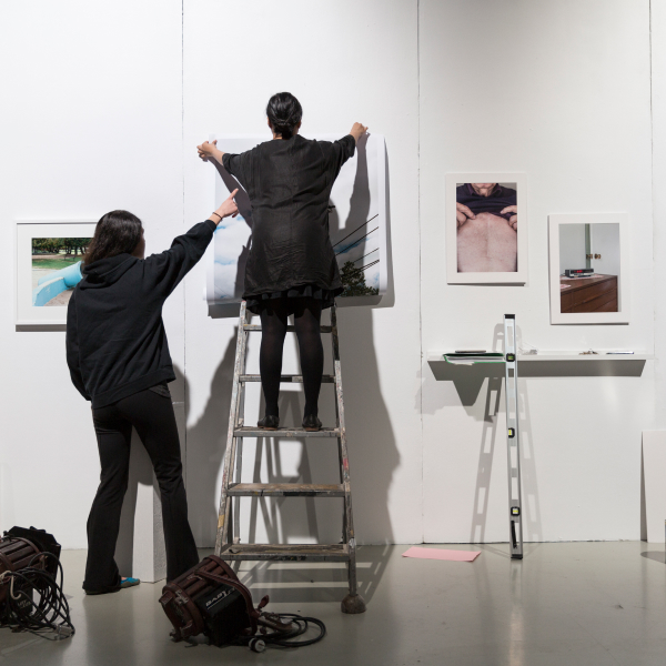 Wide shot from the back showing two people hanging a picture up on a wall. One person is on a ladder while the other props them up. There are three other pictures already on the wall.