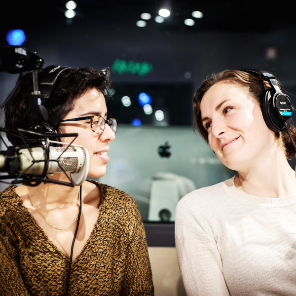 Medium close-up shot of two people in a studio looking at each other with headphones on and a mic in front of them.