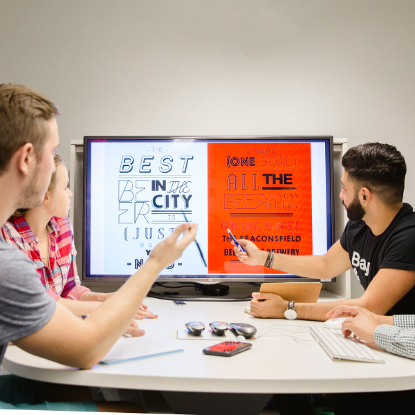 Medium-long shot of several people sitting at a round table and looking at an infographic on a TV screen. There are some writing utensils and keyboards on the table.