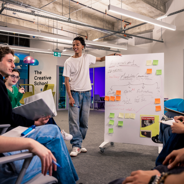 Wide shot of a man standing next to a large white board with writing and sticky notes on it. He is smiling and looking of screen. There are people sitting around him slightly off screen.
