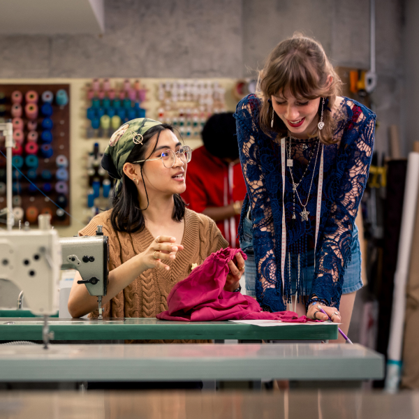 Medium long shot of two women sewing a clothing item. The person on the left is sitting down and looking towards the person on the right who is standing. They are in a sewing studio, with sewing machine, frabic, and thread rolls visible throughout the room.