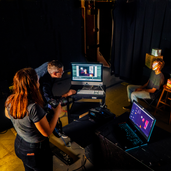 Long aerial shot of three people on a set. They are surrounded by black curtains with dim lighting, some projections, and two laptops.