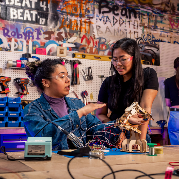 Medium shot of two women sitting at a table and talking to each other. There is a product on the table with wires and other technological parts scattered.