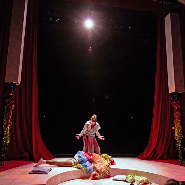 Wide shot of a woman dressed as a clown looking up. She is on stage and there is a spotlight on her.