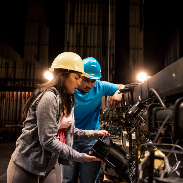 Medium-long shot of two people in hard hats operating a fly rail on a theatre stage. They are holding a lighting fixture that is attached to the rail.