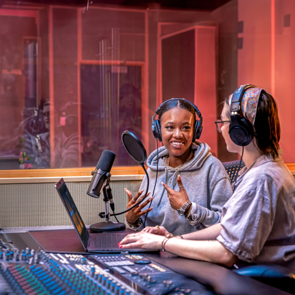 Medium shot of two people in a recording studio wearing headphones with audio control equipment in from of them.