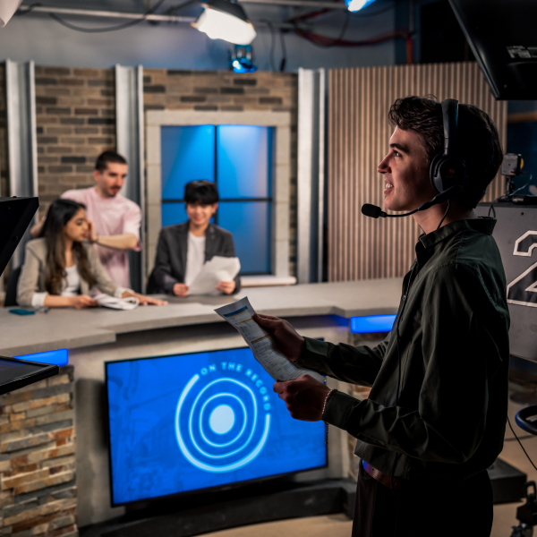 Foreground shows a medium shot of a man wearing a headset mic, holding a paper, and looking to the left. In the background, you can see three people on a news-like set.
