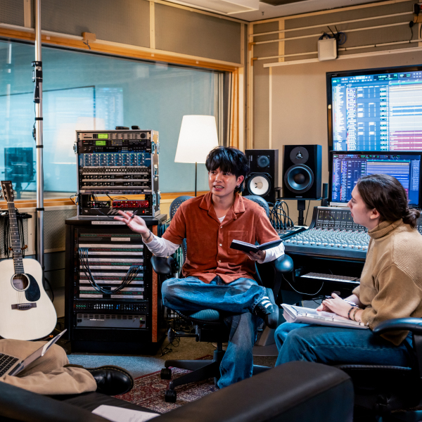 Wide shot of two people in a music recording studio talking to each other. There is various equipment in the room, including speakers, padded walls, A/V technology, couches, microphone stands, and a guitar.