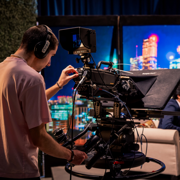 Medium-long shot of a man with headphones in a studio operating a large camera. The background shows a news-like set.