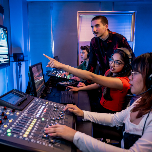 Medium shot of four people sitting in a control room. One person is point towards the large TV screen and the others look. There is various A/V tech in front of them.