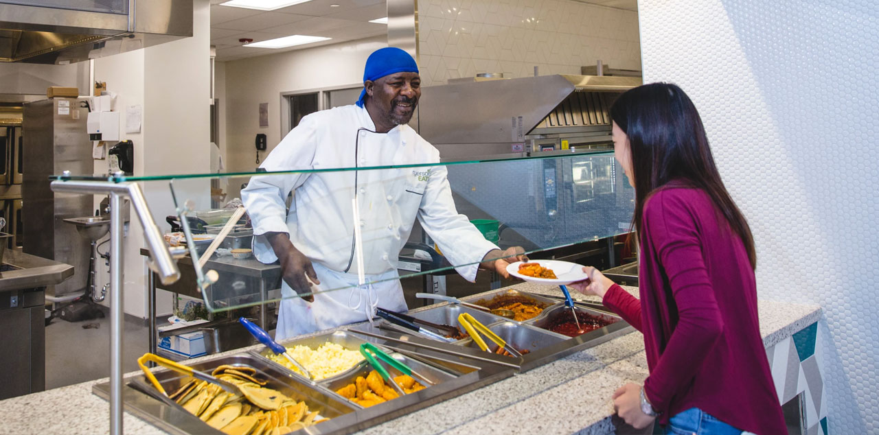 A student being served a hot meal by a chef in the new Pitman Dining Hall. A large bright sign reads Home Kitchen.