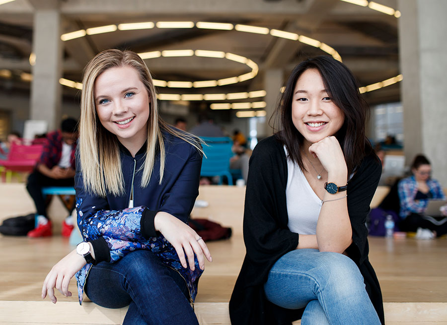 Two female students sit on the 6th floor of the SLC. They are both looking at the camera. Other students can be seen in the background.