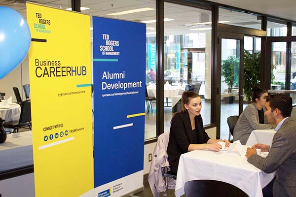 Two people sitting at table with two banners behind them that read, Business Career Hub and Alumni Development
