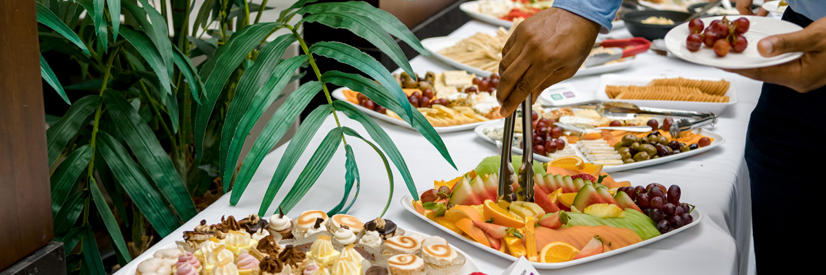 A person using tongs to take fruit from a tray amogst a large spread of food.