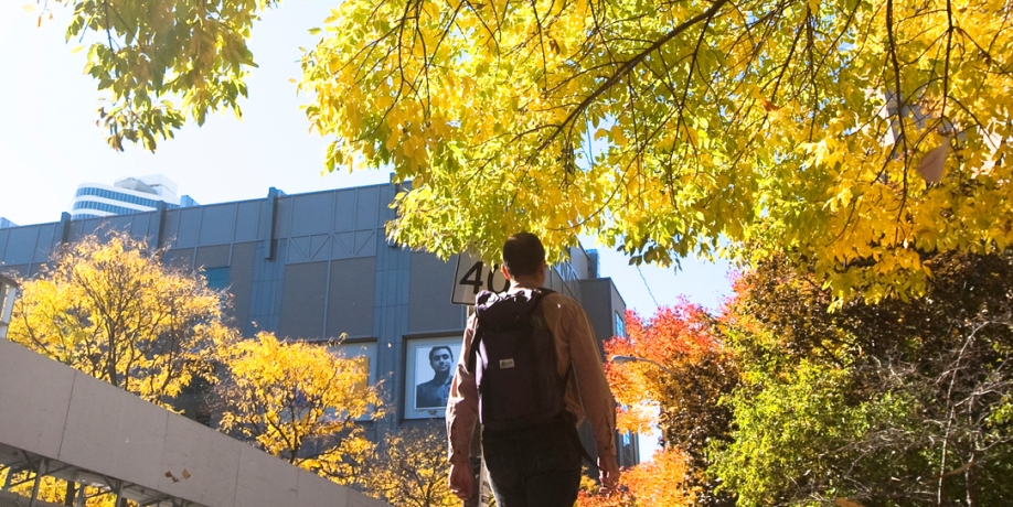 Figure with a backpack walks on campus amidst autumn foliage