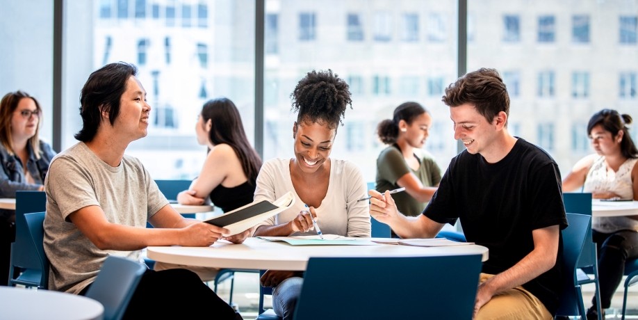 Three students smiling and working together around a table in a bright study space.