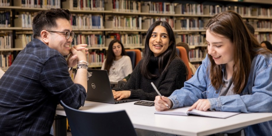 Three students smiling and studying together around a table in a library.
