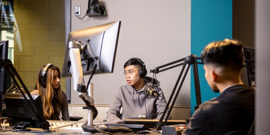Three students wearing headphones having a discussion around microphones in a recording studio.