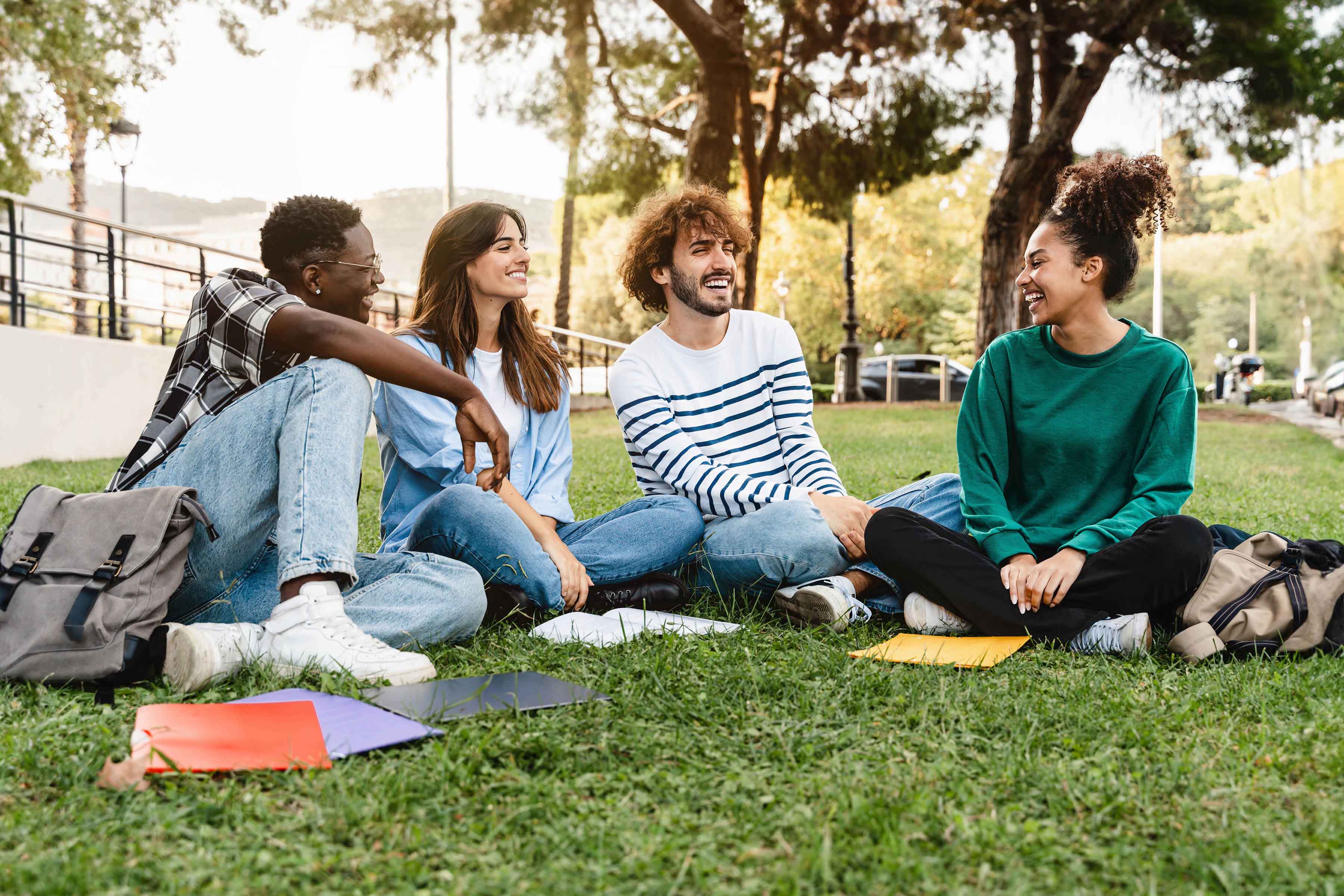 Group of four young adults sitting on the grass outdoors, smiling and talking together with notebooks and backpacks around them